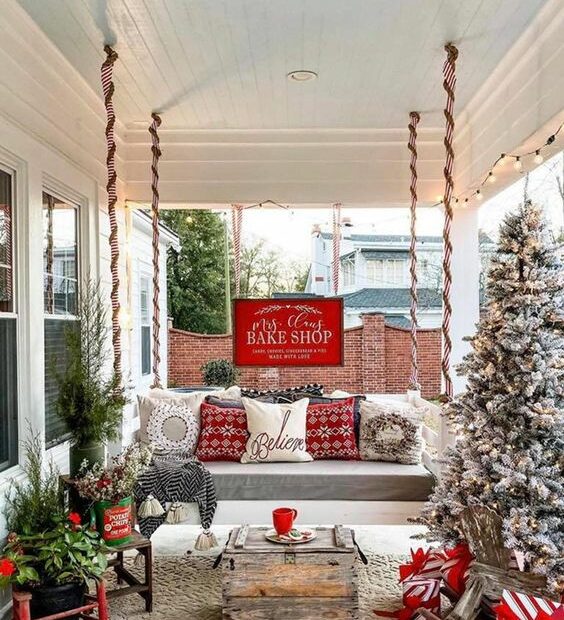 a bright rustic Christmas porch with a flocked Christmas tree with lights mini trees in buckets red pillows and a wooden table